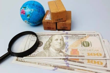 hundred dollar bills on a table with a magnifying glass, small globe, and shipping boxes